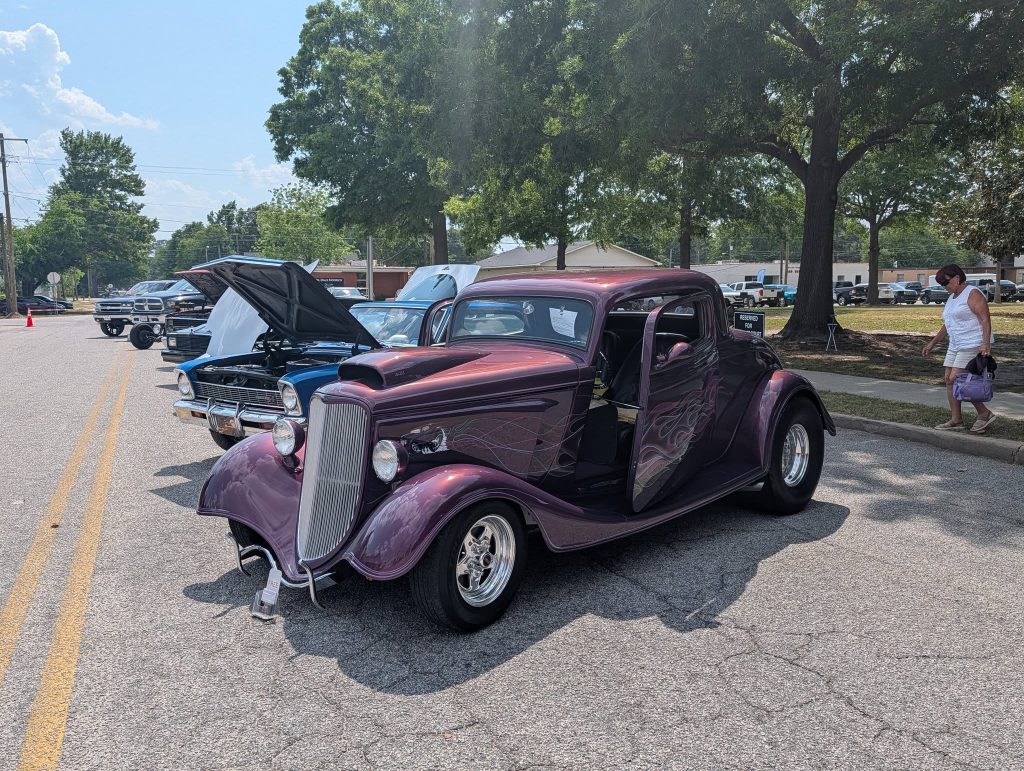 A classic purple hot rod is parked on a street at a car show, with other vintage cars lined up and people walking nearby under trees.