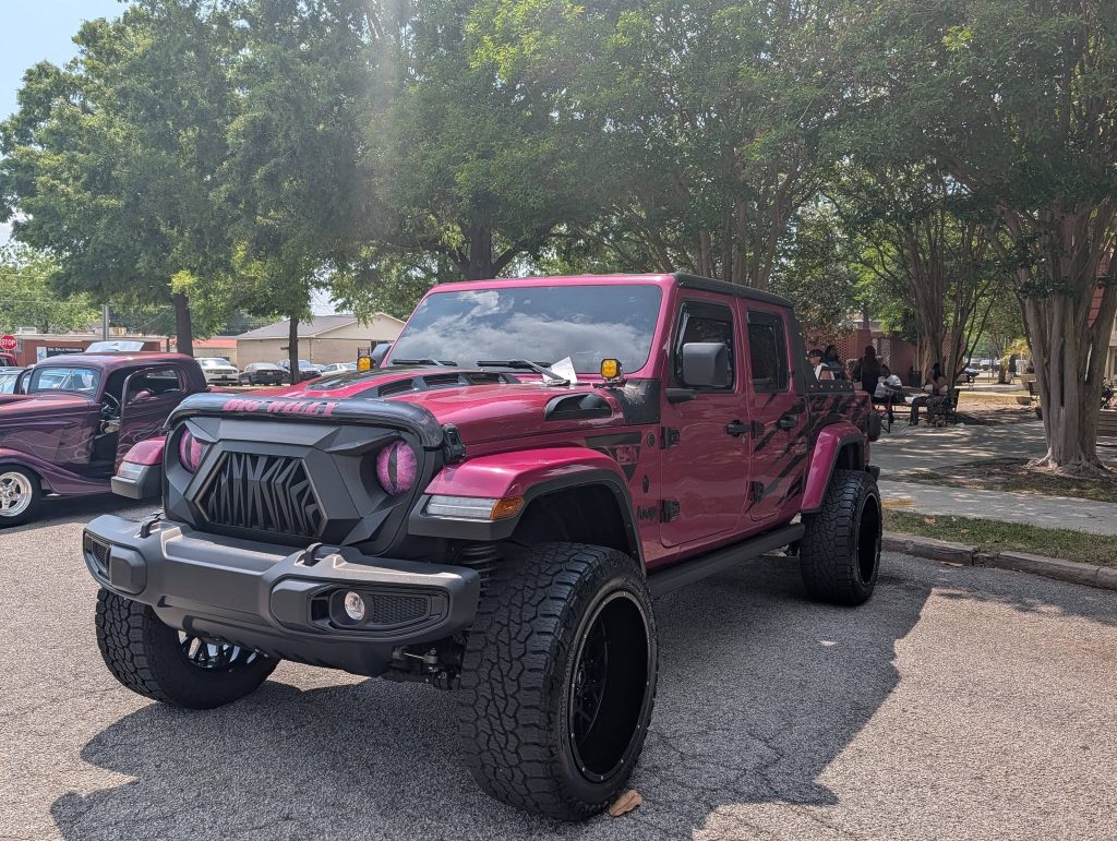 A custom magenta Jeep truck with oversized wheels and unique grille parked outdoors at a car show, with trees and people in the background.