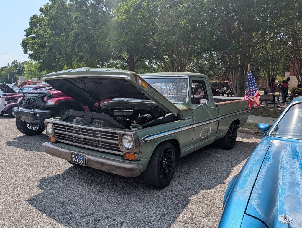 A green 1971 Ford F-100 pickup truck with its hood open, parked at a car show, and an American flag mounted in the truck bed.