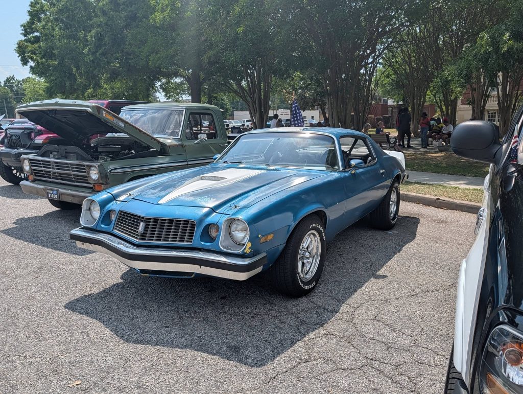 A blue vintage muscle car with white racing stripes parked at a car show, surrounded by other classic vehicles and people in the background under trees.