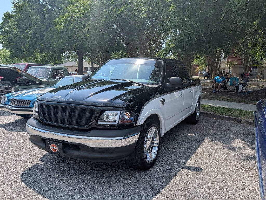 A black and white pickup truck on display at a car show, parked on a street with other vintage vehicles and people sitting under trees nearby.