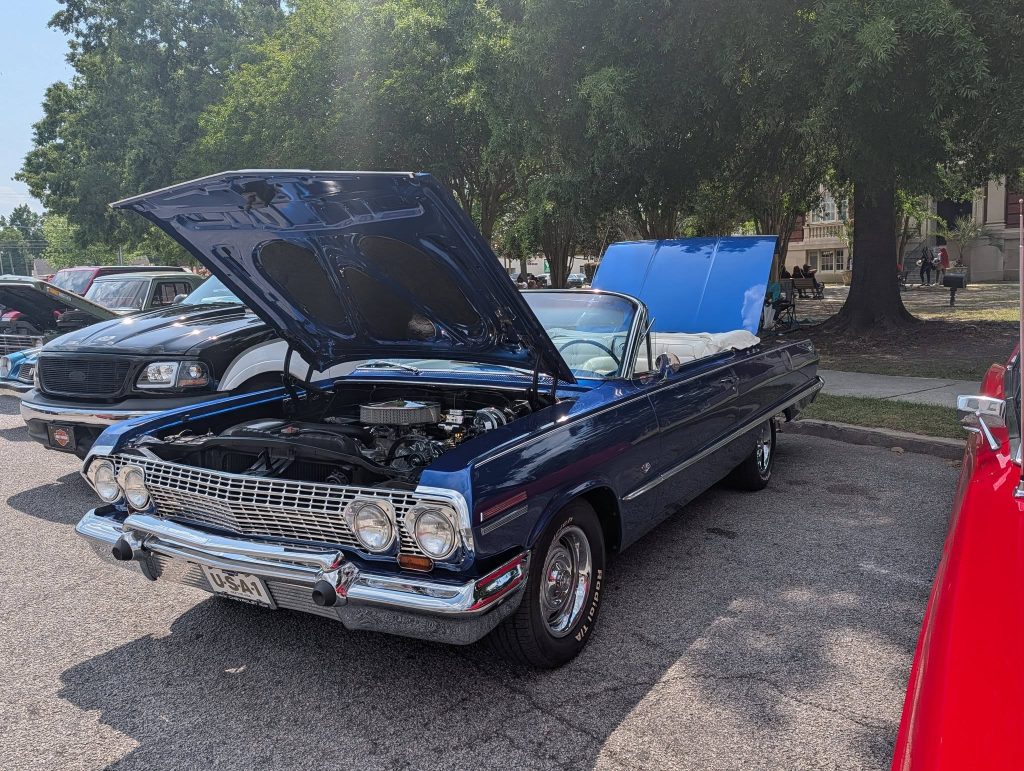 A classic blue convertible car with its hood and trunk open is parked at a car show, surrounded by other vehicles and trees.