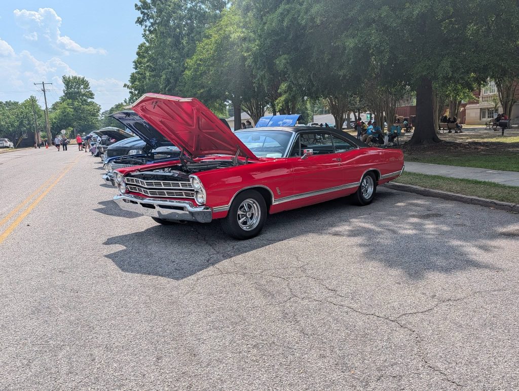 A row of classic cars with open hoods is parked on a street during a car show; people gather nearby under trees.