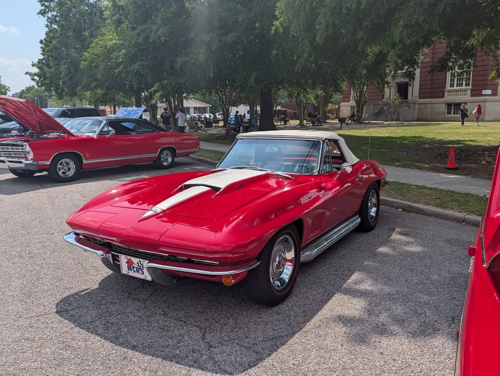 Red vintage convertible car with white hood stripe parked at a car show, surrounded by trees and other classic cars, with people in the background.