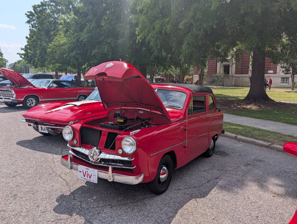 A red vintage car with its hood open at a car show, parked on a street with other classic vehicles and trees in the background.