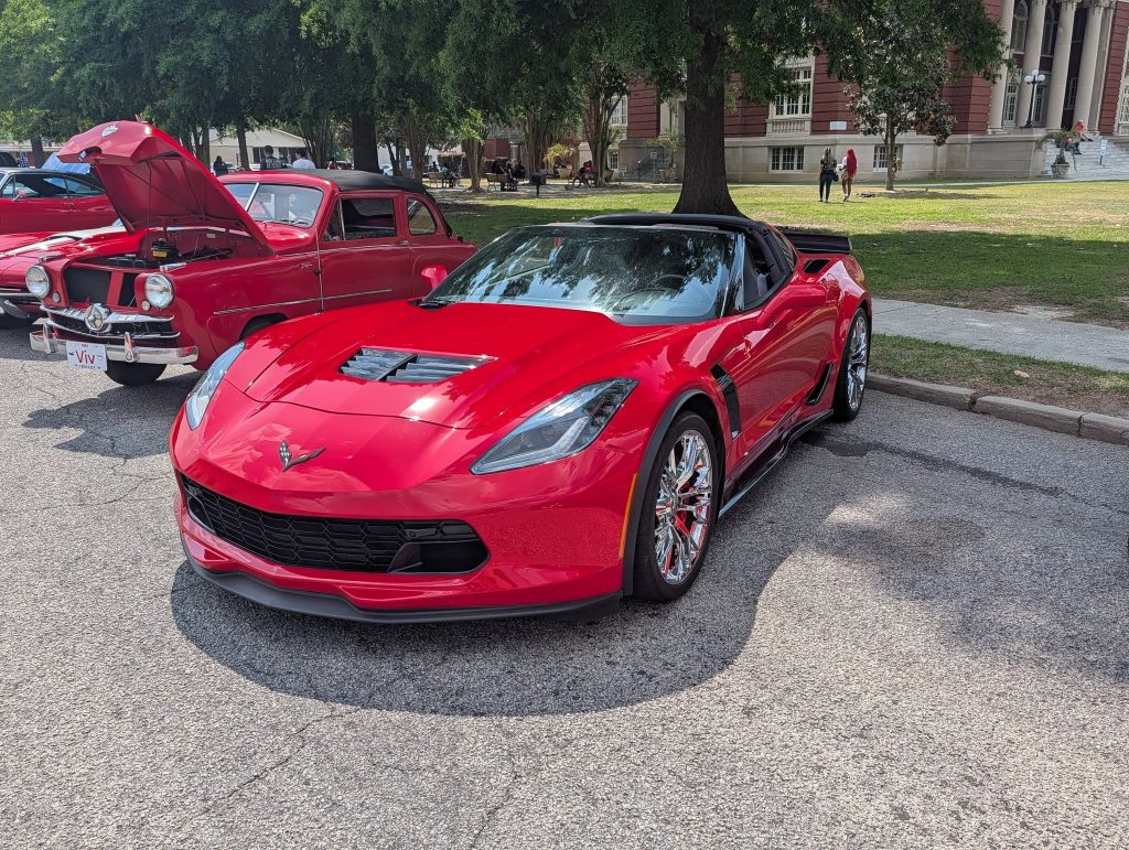 A shiny red sports car parked on the street next to a classic car with its hood open, at an outdoor car show.