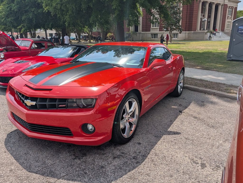 A shiny red Chevrolet Camaro with black racing stripes parked at a car show, surrounded by other classic cars and people in a park setting.