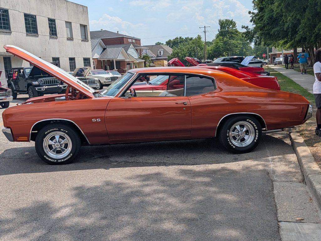A classic orange Chevrolet Chevelle SS muscle car with its hood open, parked at a car show on a sunny day.
