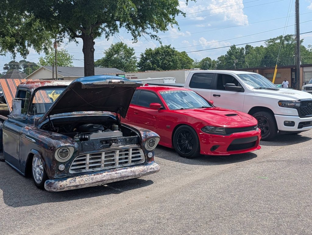 Three vehicles parked: an old pickup truck with the hood open, a red sports car, and a white pickup truck, on a sunny day.