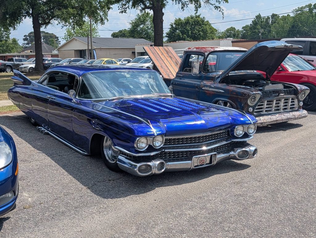 Classic blue custom lowrider car and a vintage pickup truck with an open hood parked at a car show on a sunny day.