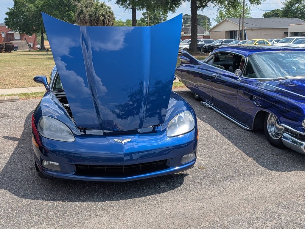 A blue sports car with its hood open, parked next to a classic car at an outdoor car show on a sunny day.