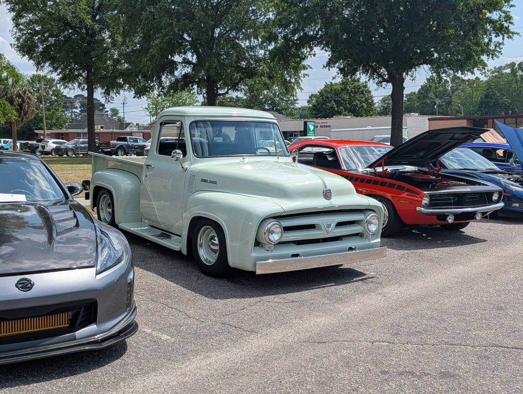 Classic cars displayed at an outdoor car show, including a light green vintage pickup truck, surrounded by other sports and muscle cars, with trees and buildings in the background.