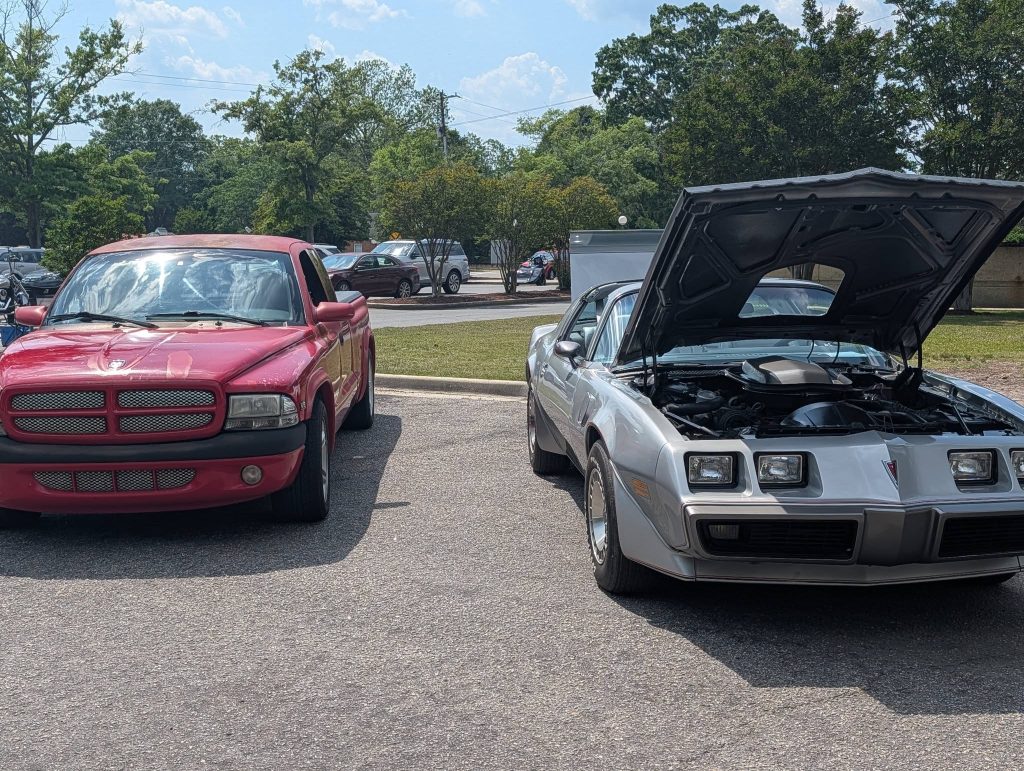 A red pickup truck parked next to a classic silver sports car with its hood open, shown in a sunlit outdoor parking lot.