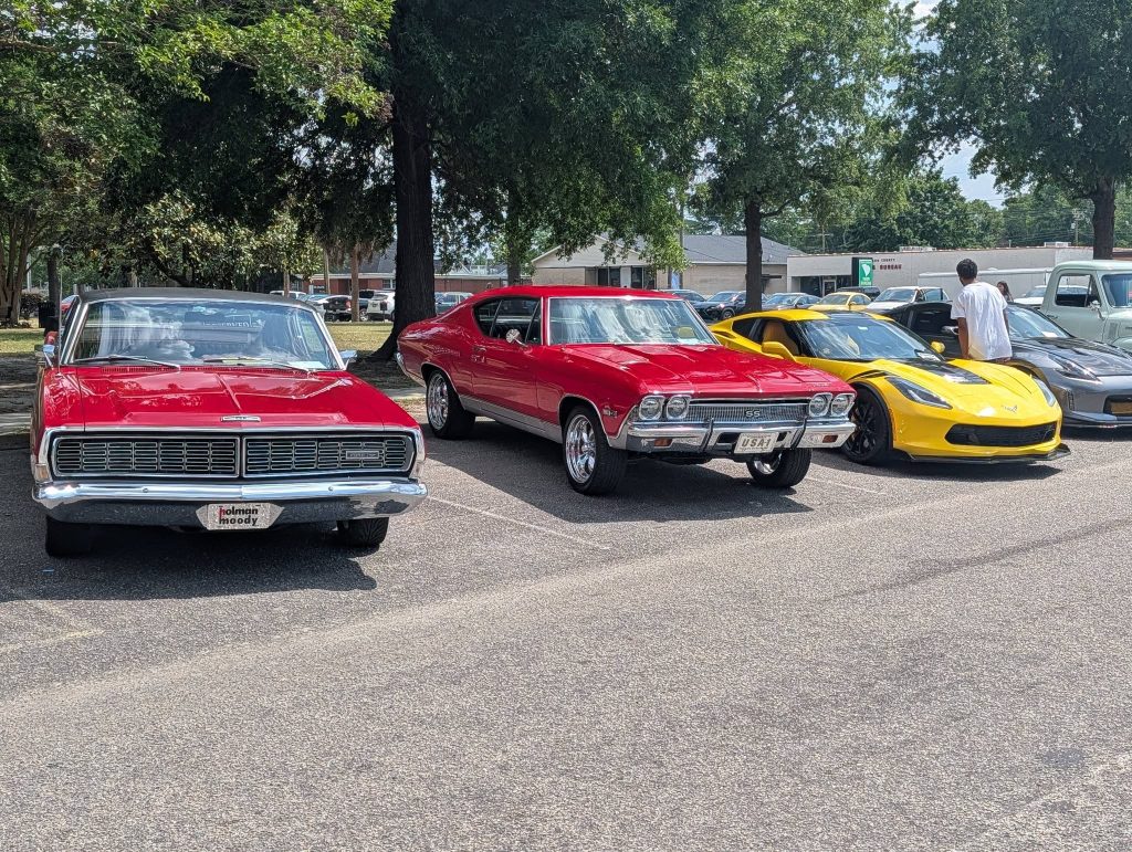 Three classic cars, two red and one yellow, parked side by side on a sunny day with trees and buildings in the background.