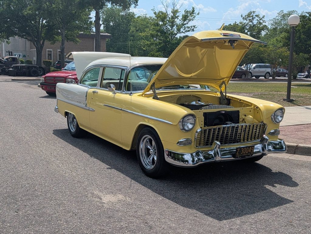 A yellow and white classic car with its hood open, parked on a street during the day. Other vehicles and trees are visible in the background.