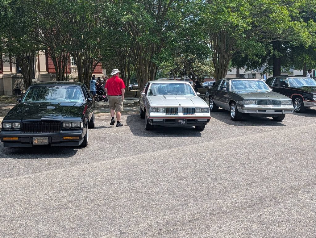 Four classic cars parked in a row at an outdoor car show, with a man in a hat and red shirt walking past them under trees.