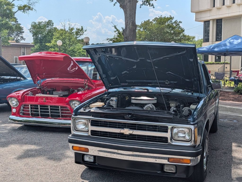 Two classic Chevrolet trucks with hoods open on display at an outdoor car show, sunny day, trees and buildings in the background.