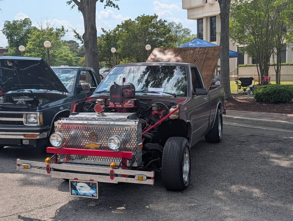 A custom pickup truck with a unique front grille and exposed engine is parked at an outdoor car show, with its hood open and another vehicle beside it.