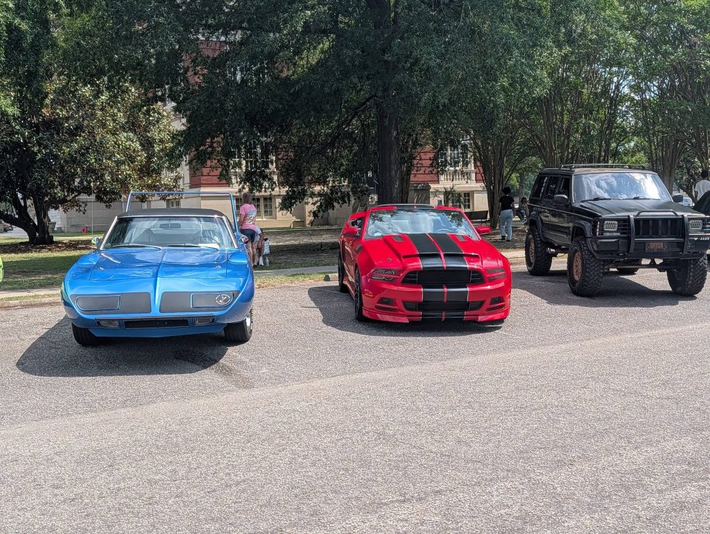 Three cars parked outdoors: a blue vintage car, a red sports car with black racing stripes, and a black off-road SUV, with trees and people in the background.