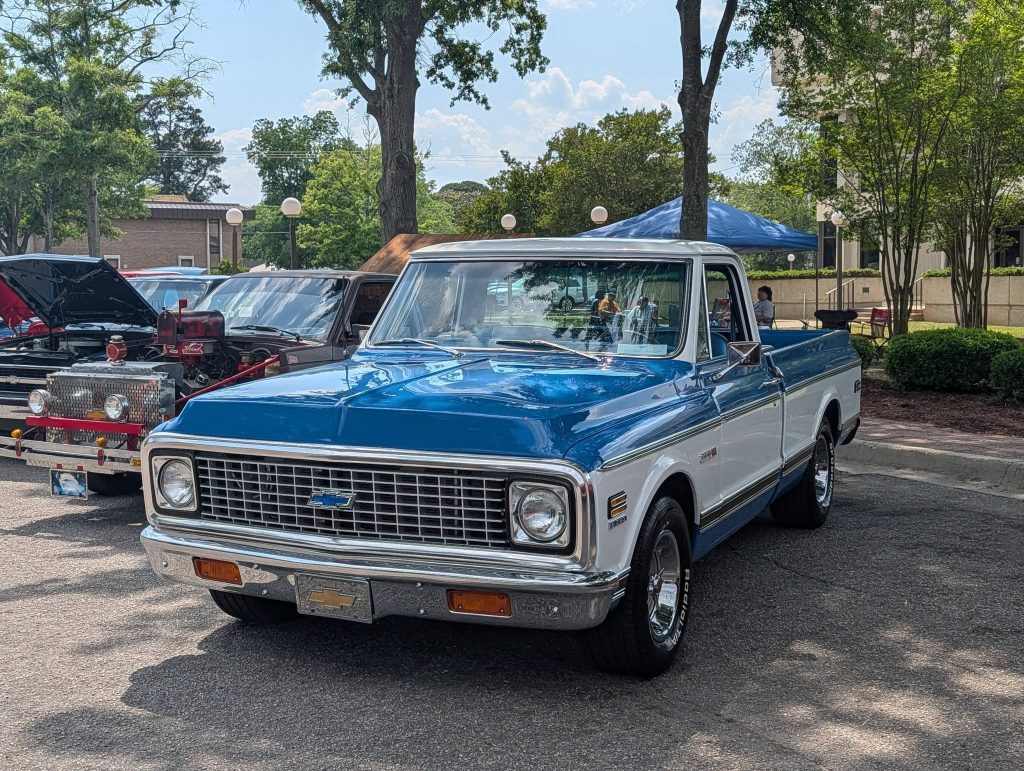 A classic blue and white Chevrolet pickup truck is parked outdoors at a car show, surrounded by other vintage vehicles and trees.