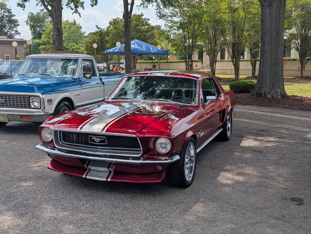 A classic red Ford Mustang with white racing stripes is parked at a car show, next to a vintage blue and white Chevrolet pickup, outdoors on a sunny day.