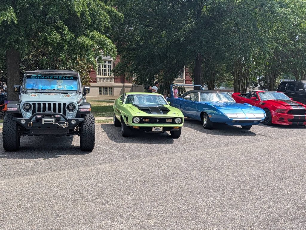 Four classic and modern cars, including a Jeep, a green muscle car, a blue sports car, and a red sports car, are parked in a lot under trees.