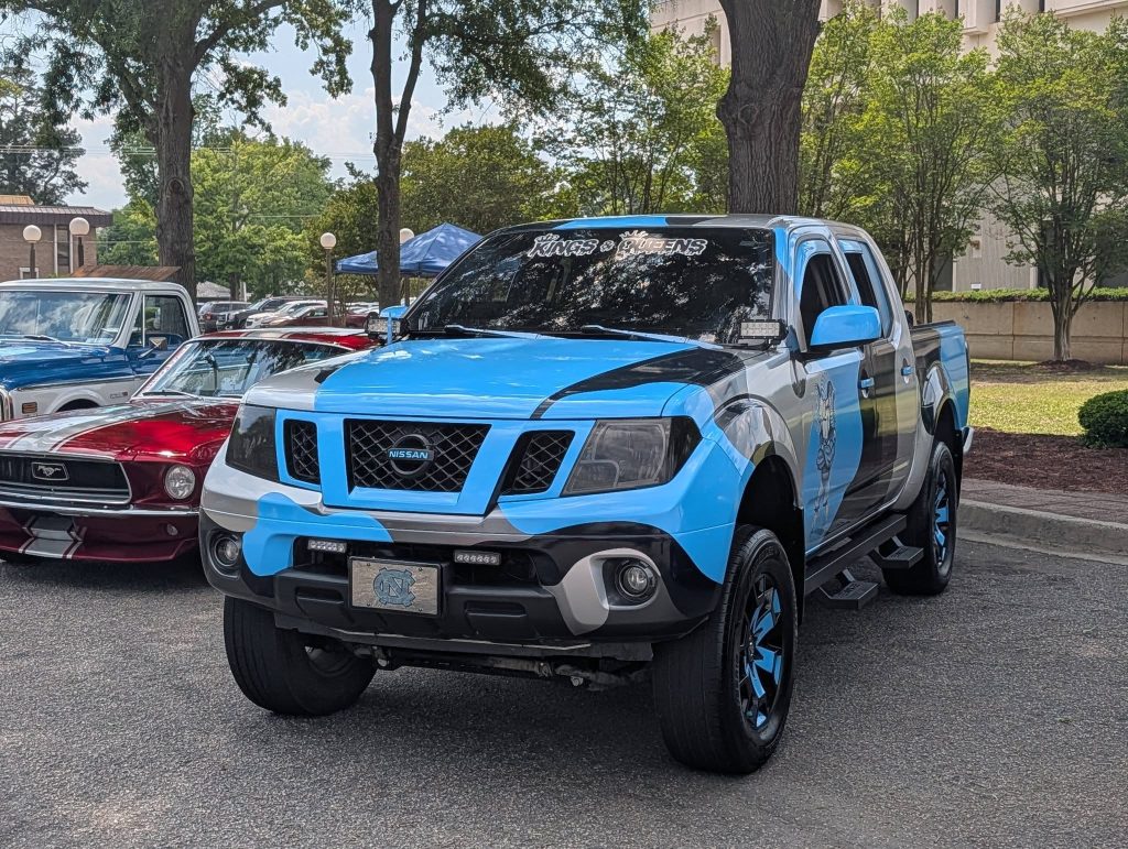 A custom Nissan pickup truck with blue, black, and white paint parked at a car show, next to classic cars, with trees and buildings in the background.