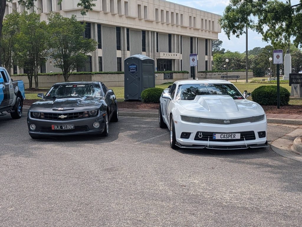 Two Chevrolet Camaros, one black and one white, parked side by side in front of a building labeled "Dillon" with trees and a portable toilet nearby.
