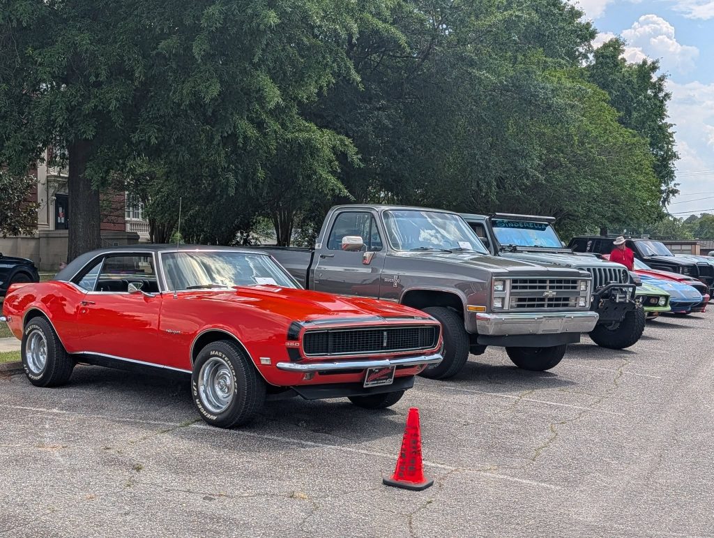 Classic cars, including a red Chevrolet Camaro and gray Chevrolet truck, parked at an outdoor car show with trees and people in the background.