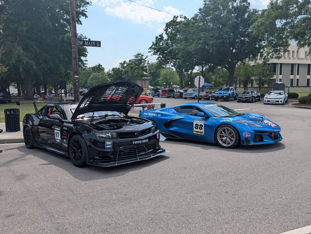 Two race cars, one black with its hood open and one blue, parked on a street at a car show with other vehicles and people in the background.