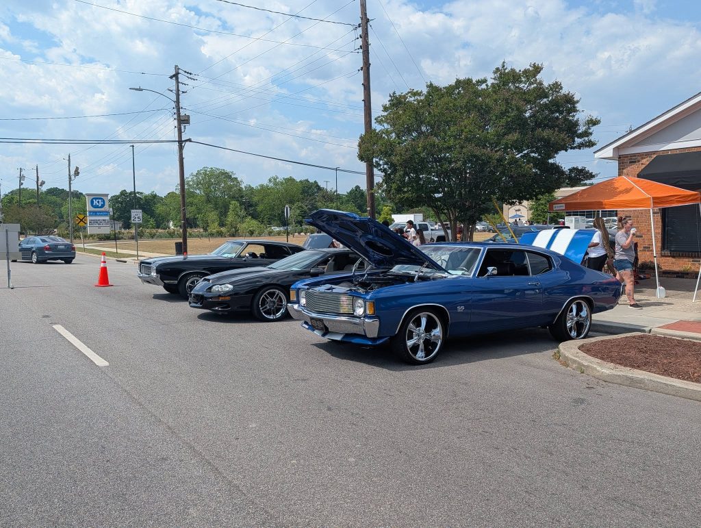 Three classic cars parked on a street, hoods open, with people standing nearby and an orange tent in the background on a sunny day.