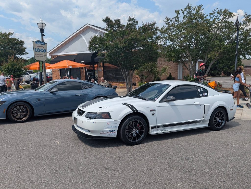 Two sports cars parked on a street in front of a building, with people walking and gathering nearby under orange canopies on a sunny day.