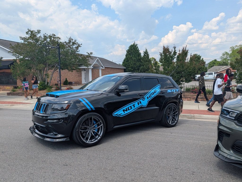 Black SUV with blue accents, "NOT A HAWK" decal, parked on a street during a daytime event, with people walking on the sidewalk nearby.