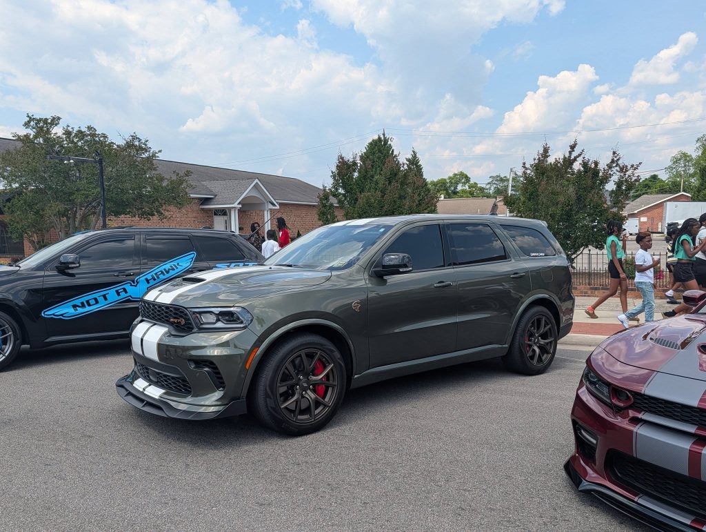A gray Dodge Durango SUV with white racing stripes parked on the street, surrounded by other cars and people walking on the sidewalk under a partly cloudy sky.