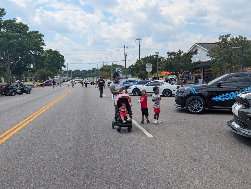 A woman pushes a stroller with a child, while two boys stand nearby on a blocked street with parked cars and people walking around.
