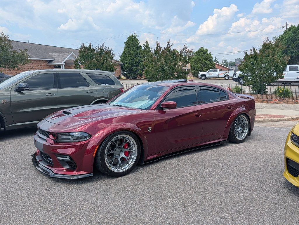 A lowered maroon Dodge Charger with custom wheels and tinted windows is parked in a lot next to other vehicles on a cloudy day.