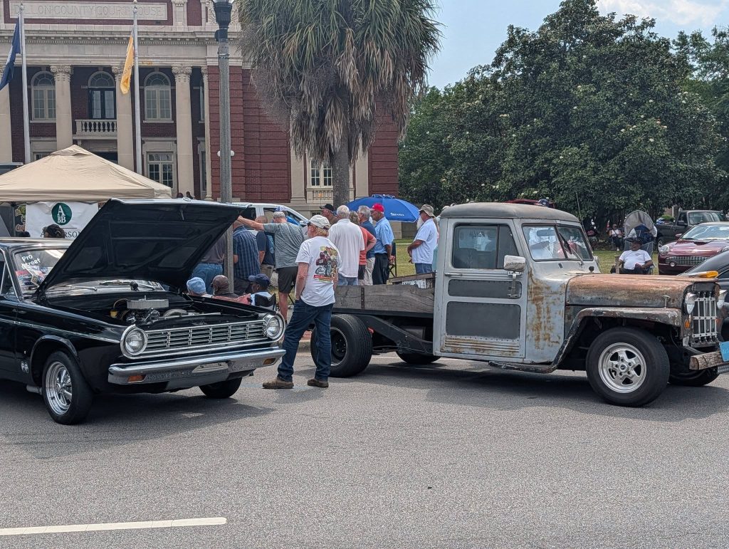 Classic car show with people viewing a black car with its hood open and an old, rusty pickup truck, outside a courthouse building.