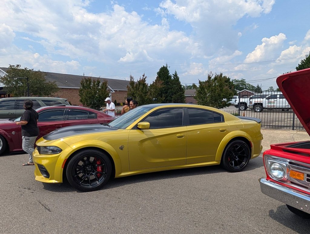A yellow Dodge Charger with black accents parked at a car show, surrounded by other vehicles and several people on a sunny day.