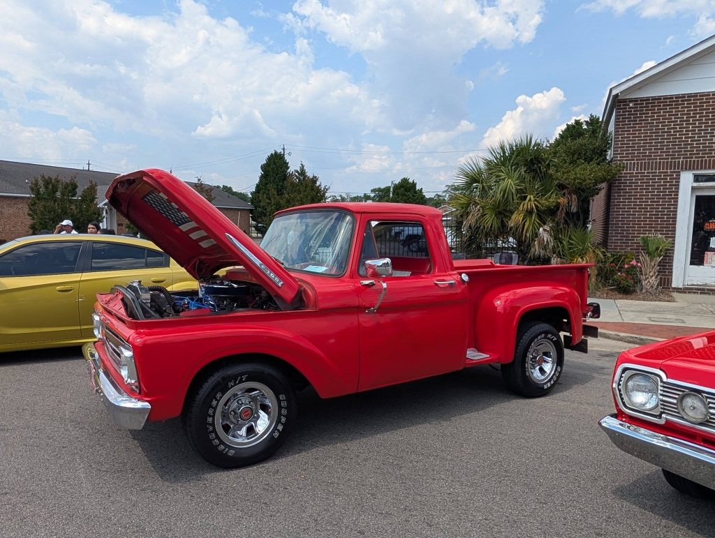 A bright red vintage pickup truck with its hood open parked on a street, with other cars and a brick building in the background.
