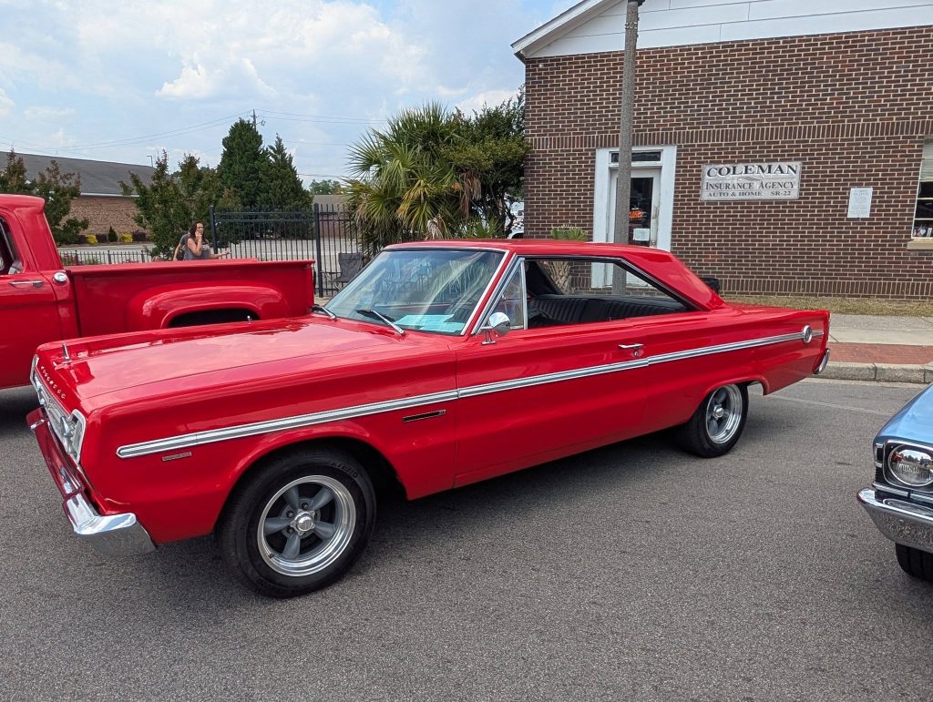 A classic red muscle car is parked on the street in front of a brick building with a sign reading "Coleman Insurance Agency."