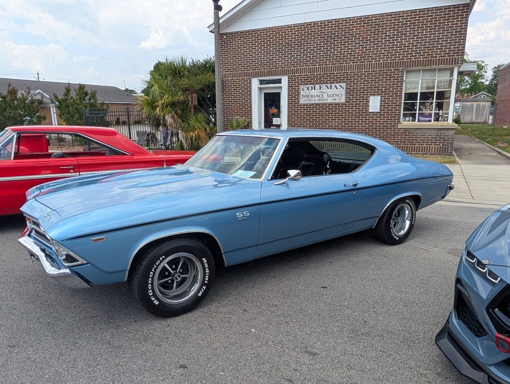 A classic blue Chevrolet Chevelle SS parked on the street in front of a brick building next to other vintage cars.