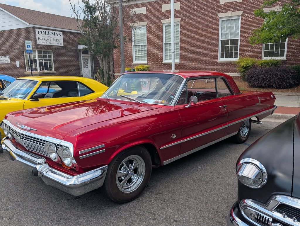 A shiny red classic car parked on the street beside other vintage vehicles, with a brick building and insurance agency visible in the background.