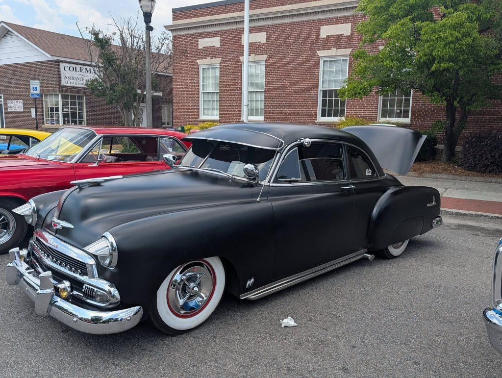 A matte black vintage Chevrolet car with chrome details and white wall tires is parked on a street near brick buildings.