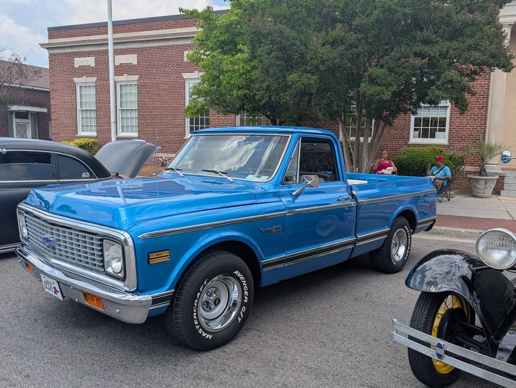 A bright blue vintage Chevrolet pickup truck parked on a street during a car show, with classic cars and people sitting nearby.