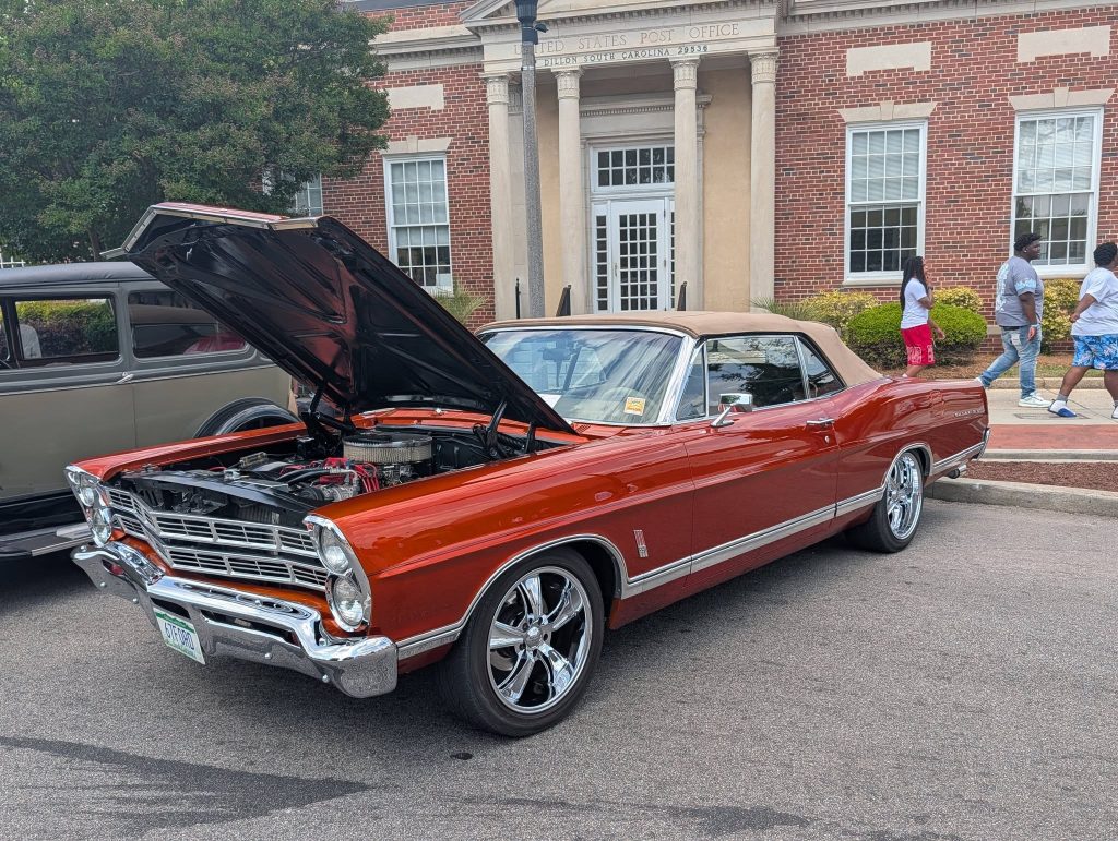 A classic red convertible car with its hood open, parked on a street in front of a brick building, people walking nearby.