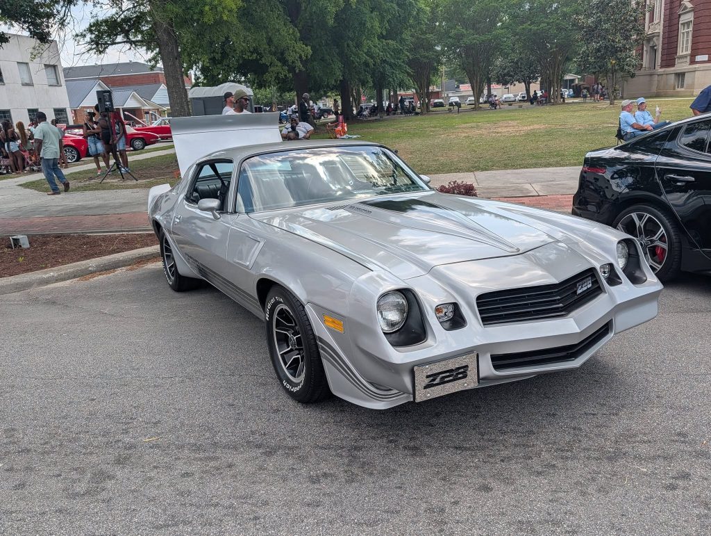 A silver classic Chevrolet Camaro Z28 parked at a car show, surrounded by people and other vehicles, with trees and buildings in the background.