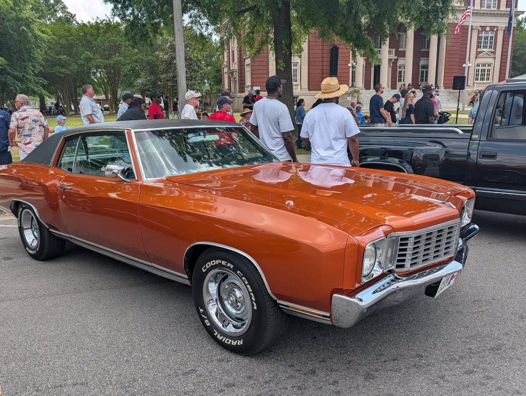 Orange classic car parked at an outdoor car show with people standing around and a historic building in the background.