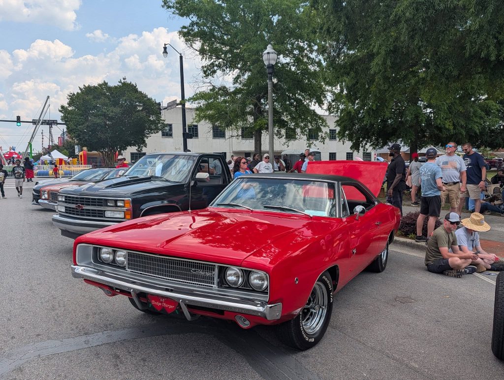 A red classic Dodge Charger with its hood open at a car show, surrounded by other vehicles and people on a sunny day.