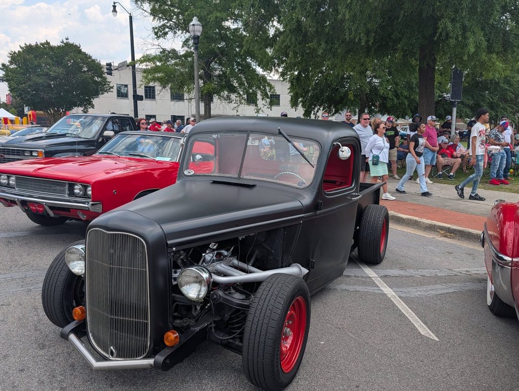 A matte black classic hot rod truck with red wheels parked at a car show, surrounded by other vintage cars and a crowd of people.