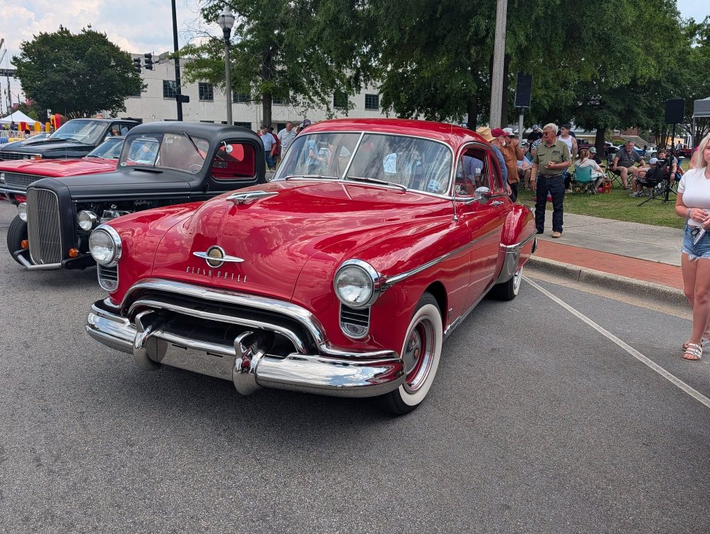 A shiny red classic Oldsmobile car parked at a car show, surrounded by people and other vintage vehicles on a city street.